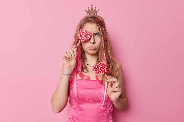 Horizontal shot of discontent young woman with long hair wears small crown and dress holds heart lollipops has sulking expression isolated over pink background. Angry princess with caramel candies