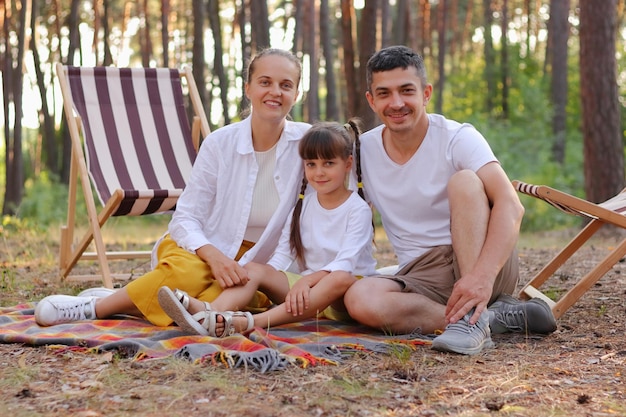 Horizontal shot of family wearing white shirts sitting on blanket in park and looking at camera with smile Caucasian parents with small daughter relaxing in forest together