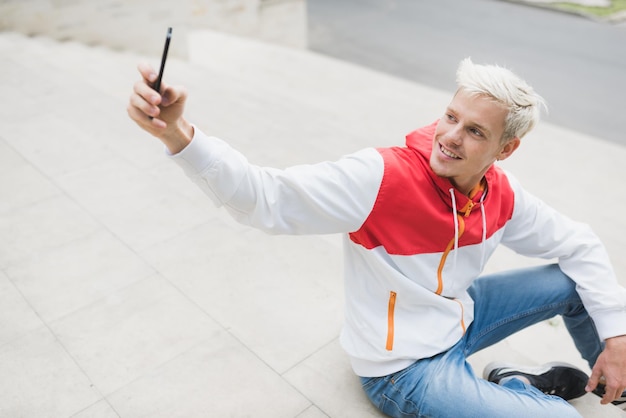 Horizontal shot of fashionable young blond hipster man with toothy smile and wearing trendy zipper hoodie take self portrait with smart phone outdoors in park People lifestyle and technology concept