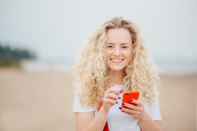 Horizontal shot of good looking young blonde female has curly hair, holds orange smart phone.