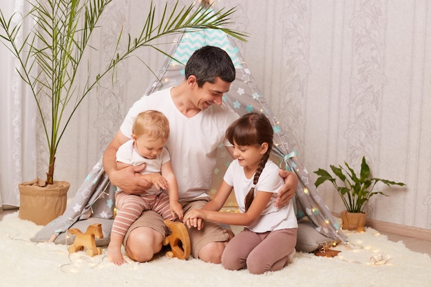 Horizontal shot of handsome man wearing white t shirt sitting on floor with her daughters and having fun playing together hugging and smiling happy family at home