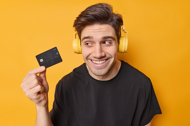 Horizontal shot of happy brunet man holds credit card glad to get money on his bank account wears stereo headphones on ears dressed casually isolated over yellow bacground thinks what to buy