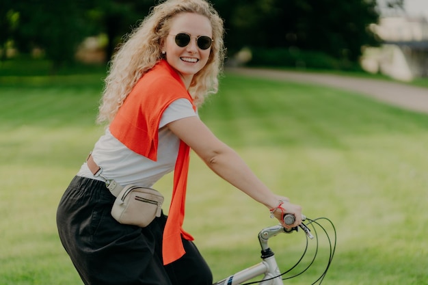 Horizontal shot of happy curly woman rides bike, keeps hands on handlebars, looks active and refreshed, dressed casually, wears shades, poses against green nature background. Spare time concept