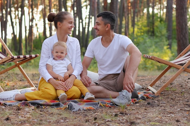 Horizontal shot of happy family enjoying moment while sitting on blanket in wood with infant daughter and looking at each other with love and gentle parents with small daughter relaxing in forest