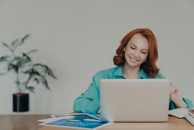 Horizontal shot of happy ginger woman freelancer works on laptop computer at home studies online uses software surfs information in internet sits at desktop smiles positively works remotely