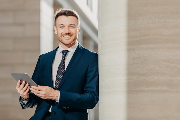 Horizontal shot of happy male manager dressed in black formal suit uses modern tablet computer for making financial report focused into distance with happy expression People and business concept