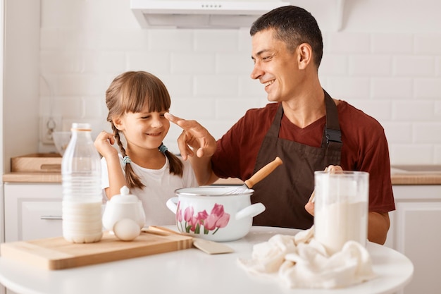 Horizontal shot of happy smiling brunette father wearing burgundy t shirt baking together with his female child in kitchen while sitting at table, father smeared the child's nose with flour.