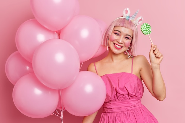 Horizontal shot of happy young Asian woman celebrates birthday holds inflated balloons makes decorations for party holds sweet lollipop wears fashionable dress 