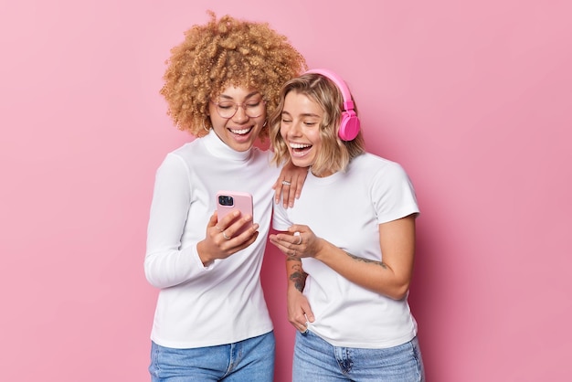 Horizontal shot of happy young women feel cheerful look gladfully at smartphone screen listen music via stereo headphones dressed in casual clothes use modern technologies isolated over pink wall