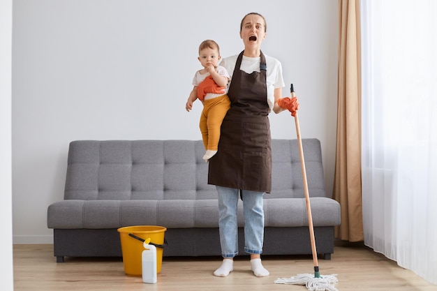 Horizontal shot of little baby girl in young mother hands desperate woman cleaning at home tired exhausted mom mopping floor tidying together in living room