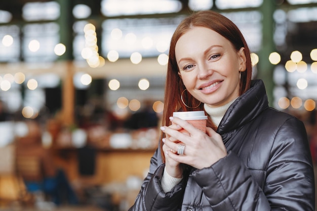 Horizontal shot of lovely woman with toothy smile holds paper cup of drink enjoys aromatic coffee wears fashionable jacket poses over blurred background with copy space Street style concept