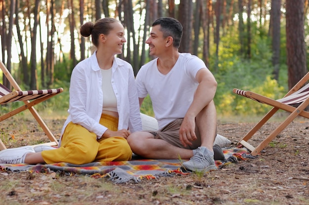 Horizontal shot of loving couple sitting in the park on the ground and looking at each other with love expressing romantic feelings wearing casual clothing having rest in the wood