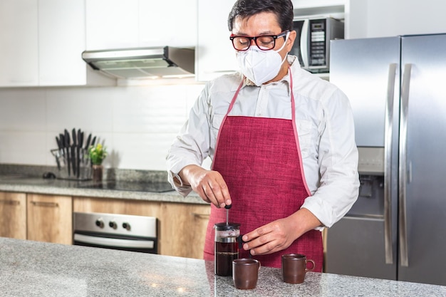 Horizontal shot of man in red apron and mask making fresh coffee in a French press next to two espresso cups in a kitchen