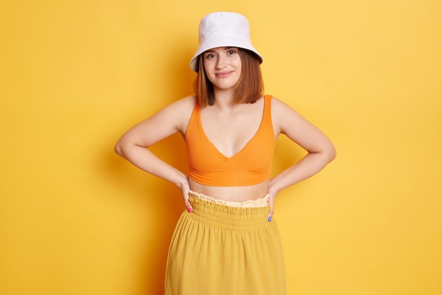 Horizontal shot of optimistic woman wearing top and panama standing against yellow wall posing with hands on hips and looking at camera expressing positive emotions