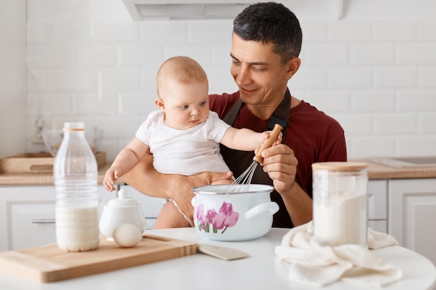 Horizontal shot of positive good looking joyful dad sitting at table with toddler daughter and cooking together in kitchen mixing dough for bakery with tiny baby's hand