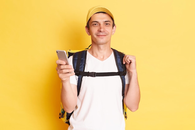 Horizontal shot of positive optimistic delivery man in yellow cap and tshirt with thermal backpack holding mobile phone looking at at camera standing with phone isolated on yellow background