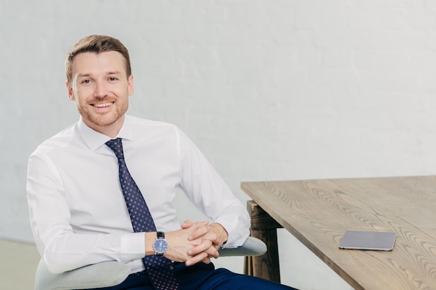 Horizontal shot of prosperous male entrepreneur in formal clothes holds hands together has positive expression sits at wooden table with touch pad waits for business partners to have meeting