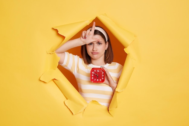 Horizontal shot of sad Caucasian woman wearing hair band and striped t shirt standing in yellow paper hole holding in hand red alarm clock and showing looser gesture