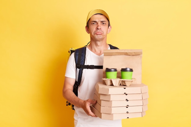 Horizontal shot of sad delivery man wearing white t shirt holding food order pizza boxes and coffee looking at camera with pout lips and expressing sadness posing isolated over yellow background