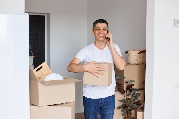 Horizontal shot of satisfied brunette man wearing white t shirt standing with cardboard box in hands and talking on smart phone looking at camera with positive facial expression