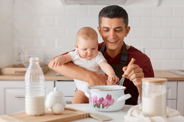 Horizontal shot of satisfied father in apron sitting at table with baby daughter and cooking together in kitchen mixing dough for homemade pastry expressing happiness