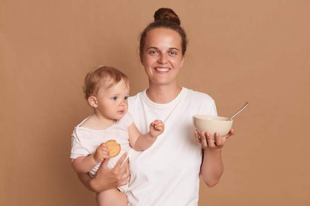 Horizontal shot of satisfied mother with bun hairstyle wearing white t shirt posing baby daughter in hands looking at camera with toothy smile holding plate with food for feeding child