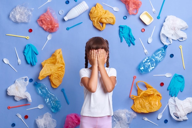 Horizontal shot of scared little girl in white Tshirt picked plastic litter isolated on blue wall covering eyes with palms hiding her face posing surrounded with much garbage