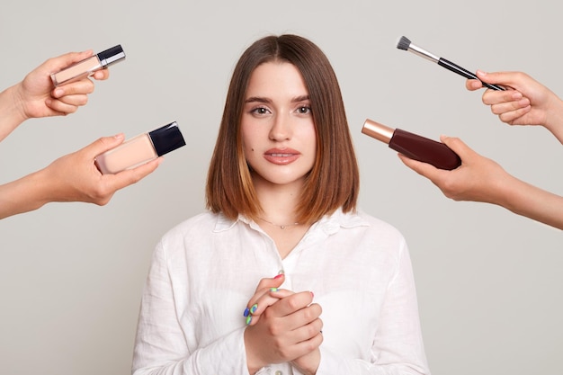 Horizontal shot of serious calm dark haired Caucasian woman standing hands together looking at camera female with fashionable makeup surrounded with stylist's hands with cosmetics