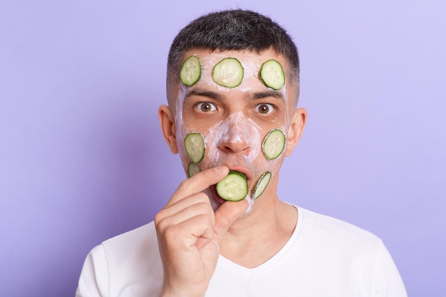 Horizontal shot of shocked man looking at camera with big eyes wearing white t shirt applying mask covering mouth with cucumber slices isolated over purple background