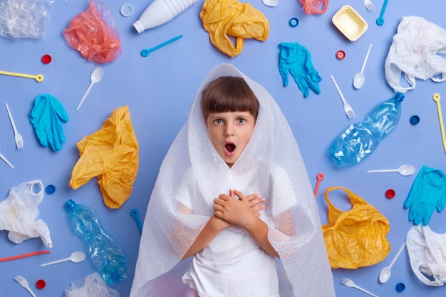 Horizontal shot of shocked stressed little girl posing against blue wall and much plastic garbage around being astonished of planet pollution looking at camera with big eyes