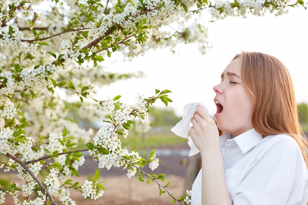 Horizontal shot of sick young woman sneezes in handkerchief, dressed in elegant shirt, has allergy to blossom, dressed in elegant shirt