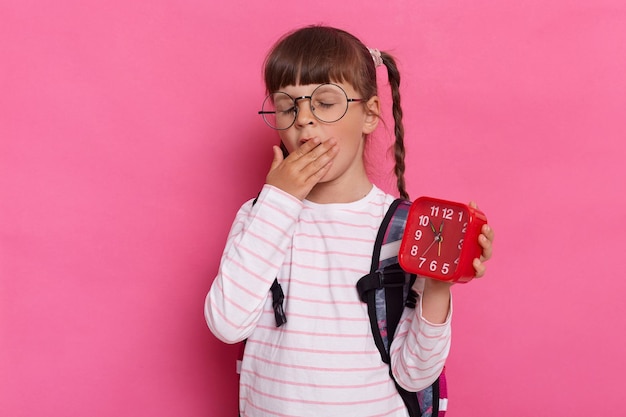 Horizontal shot of sleepy tired little schoolgirl wearing shirt and eyeglasses standing with backpack isolated over pink background holding red alarm clock yawning covering mouth with palm