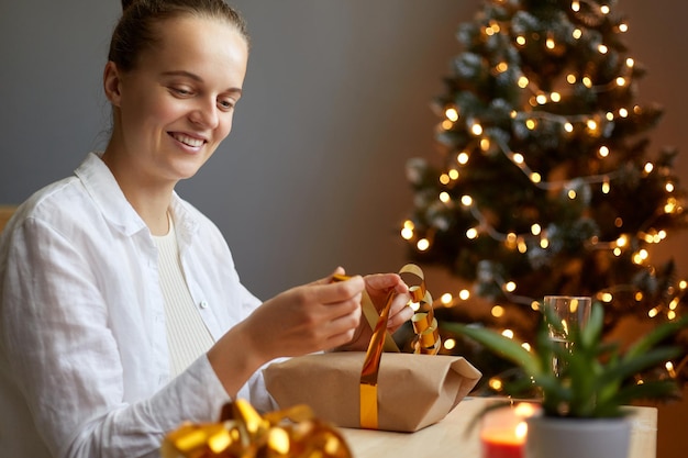 Horizontal shot of smiling adorable female packing christmas gift with golden bow on background of christmas tree with light wrapping presents for her family and friends for new year holidays