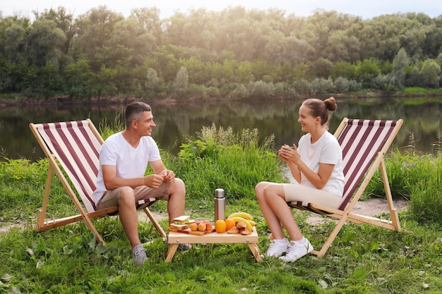 Horizontal shot of smiling happy husband and wife sitting on deck chair and drinking coffee near the river family enjoying weekend together on nature with beautiful view