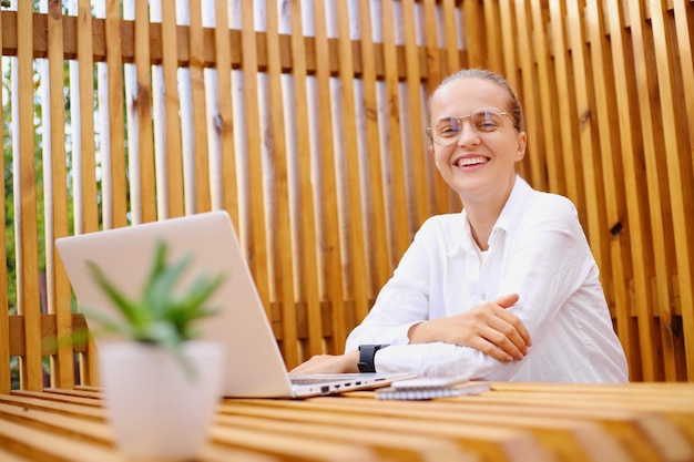 Horizontal shot of smiling happy woman using laptop working on notebook sitting at table with positive facial expression wearing white shirt looking at camera