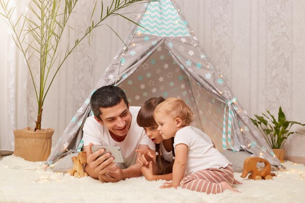 Horizontal shot of smiling man in white t shirt laying on floor with children holding smart phone in hands kids looking at mobile phone display with astonished expressions