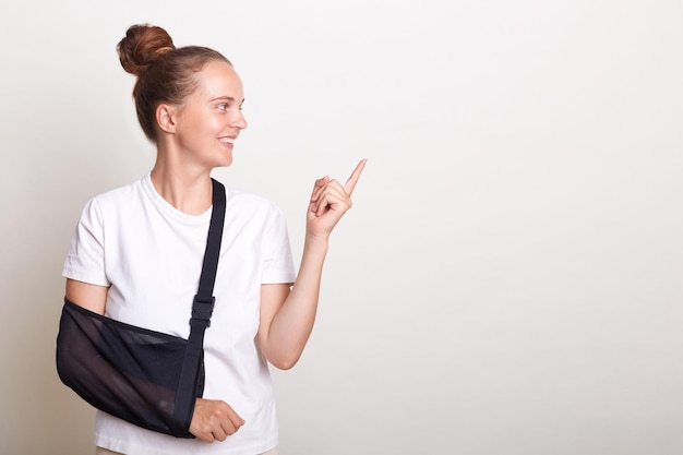 Horizontal shot of smiling positive woman with bun hairstyle wearing casual t shirt posing isolated over white background female with broken arm in splint pointing away at advertisement area