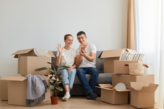 Horizontal shot of smiling young family buying new flat and posing surrounded with boxes with personal pile sitting on sofa and having video call waving hands to phone camera