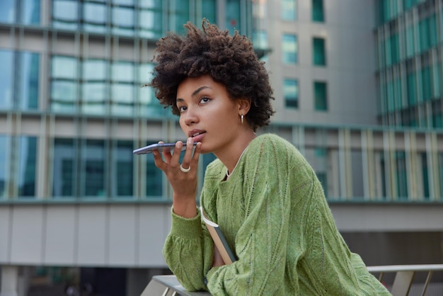 Horizontal shot of thoughtful curly haired woman uses cellphone for communication over speaker records voice message dressed casually poses against modern city building Modern technologies