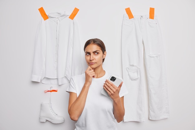 Horizontal shot of thoughtful dark haired young woman considers something with serious expression holds mobile phone poses against white background plastered items of clothes thinks what to wear