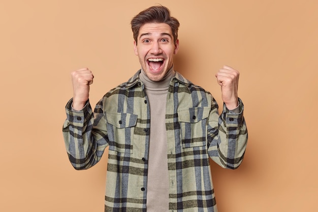 Horizontal shot of upbeat handsome man cleches fists celebrates achievement exclaims loudly wears casual checkered shirt feels like winner isolated over beige background Yes finally success