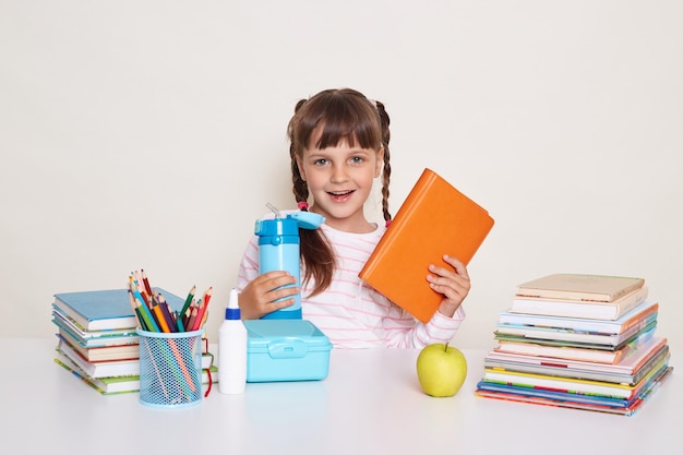 Horizontal shot of winsome positive little schoolgirl with dark hair and braids holding book and blue water bottle sitting at table surrounded with books against white wall