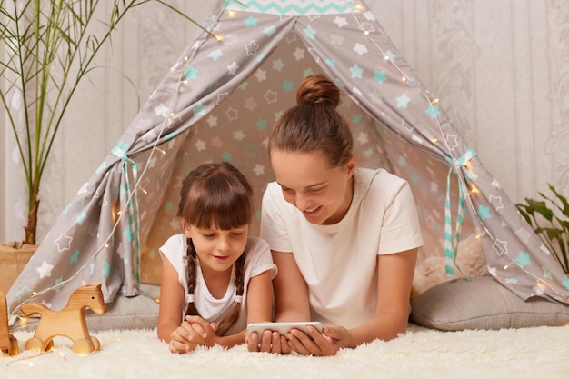 Horizontal shot of woman and her daughter laying in peetee tent and watching cartoons on mobile phone spending time together at home having fun in wigwam