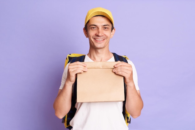 Horizontal shot of young adult handsome Caucasian delivery man holding paper bag with takeaway food very happy looking at camera with positive expression