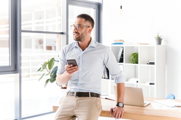 Horizontal stylish man wearing glasses sitting on table in office and looking through big window, while using mobile phone