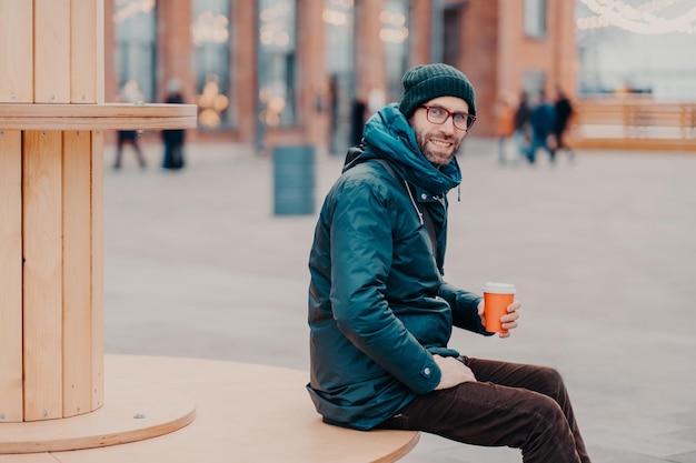 Horizontal view of cheerful European man with thick bristle wears hat and jacket holds disposable cup of coffee breathes fresh air poses at street Male model enjoys fresh beverage outside