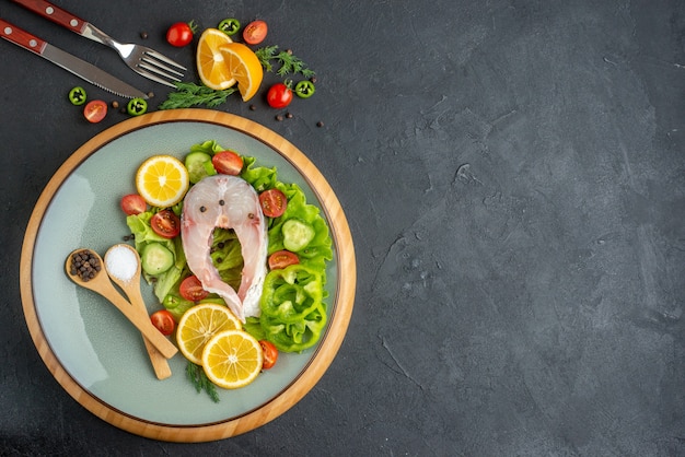 Horizontal view of raw fish and fresh chopped vegetables lemon slices spices on a gray plate and cutlery set on the right side on black distressed surface