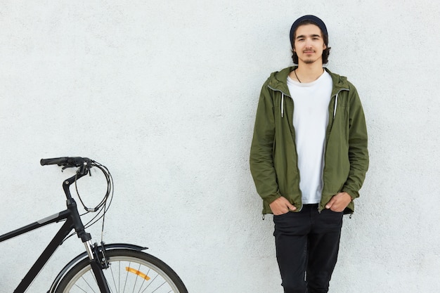 Horizontal view of stylish man keep hands in pockets of black trousers wears green jacket, stands near his bicycle, likes cycling