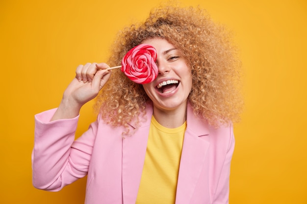 Photo horizotal shot of curly happy woman covers eye with tasty sweet candy has fun laughs happily dressed in formal clothes isolated over vivid yellow wall. lovely glad female holds lollipop