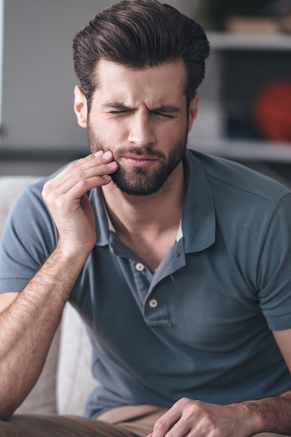 Horrible toothache. Frustrated young man touching his cheek and keeping eyes closed while sitting on the couch at home